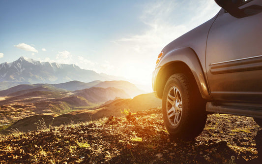 Front of a dark SUV parked on rocky terrain at sunrise or sunset, with scenic mountain ranges in the background.
